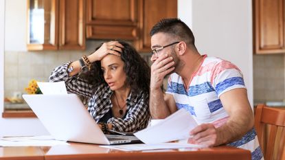 A couple looks surprised and stressed as they look at paperwork in their kitchen.