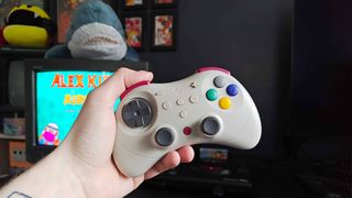 Hand holding Gulikit Elves 2 Pro controller with Sony Trinitron CRT TV and Sega Mega Drive in backdrop with Alex Kidd in Miracle World title on screen.