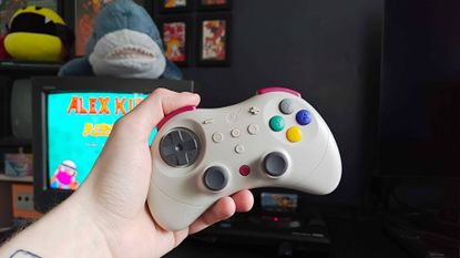 Hand holding Gulikit Elves 2 Pro controller with Sony Trinitron CRT TV and Sega Mega Drive in backdrop with Alex Kidd in Miracle World title on screen.