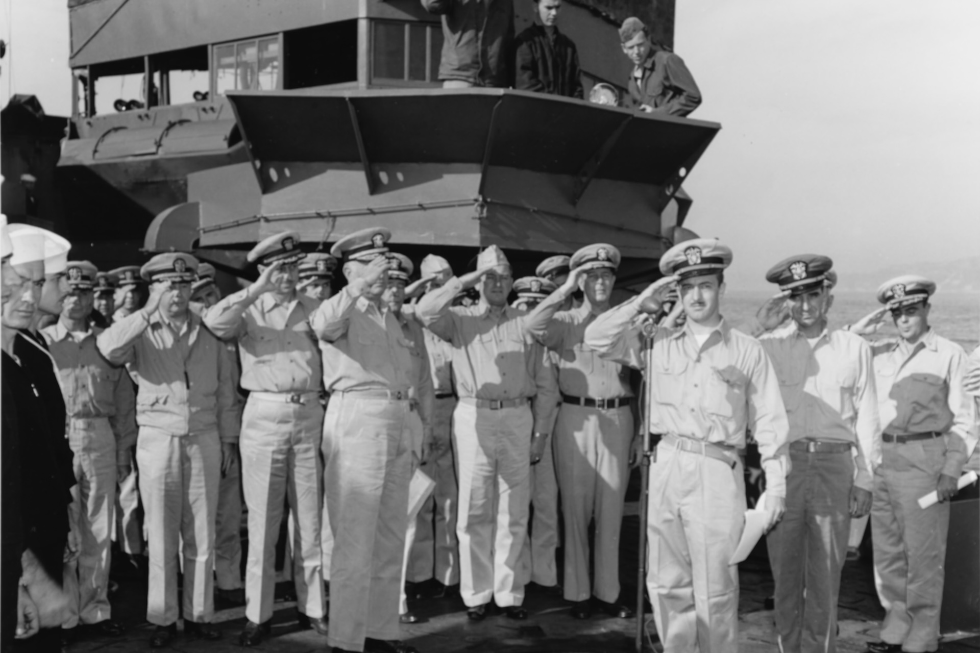 A black and white photo of over a dozen sailors saluting in front of a war ship