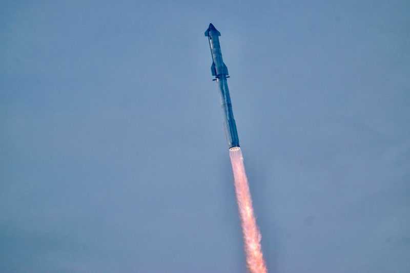 Eine große silberne Rakete fliegt über einer großen Feuersäule durch den blauen Himmel