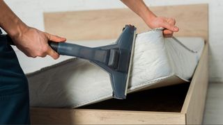 A person lifting up the corner of a mattress, holding a mattress vacuum head to the side of the mattress.