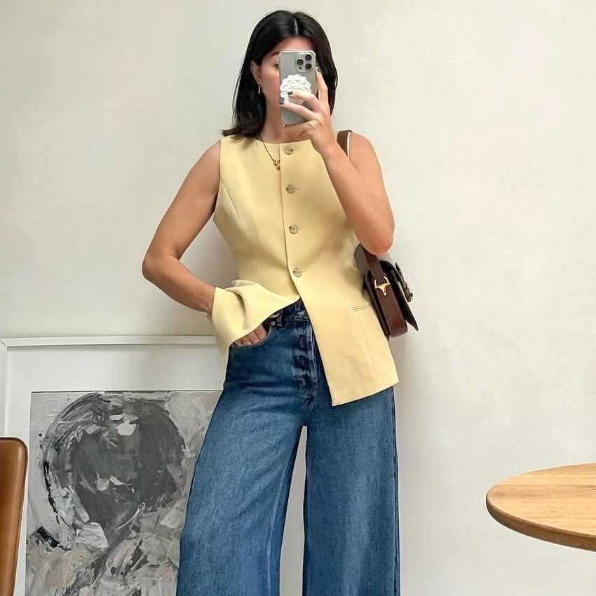 British influencer Francesca Saffari poses for a mirror selfie in her home wearing a summer outfit with butter-yellow button vest top, brown leather bag, and wide-leg jeans