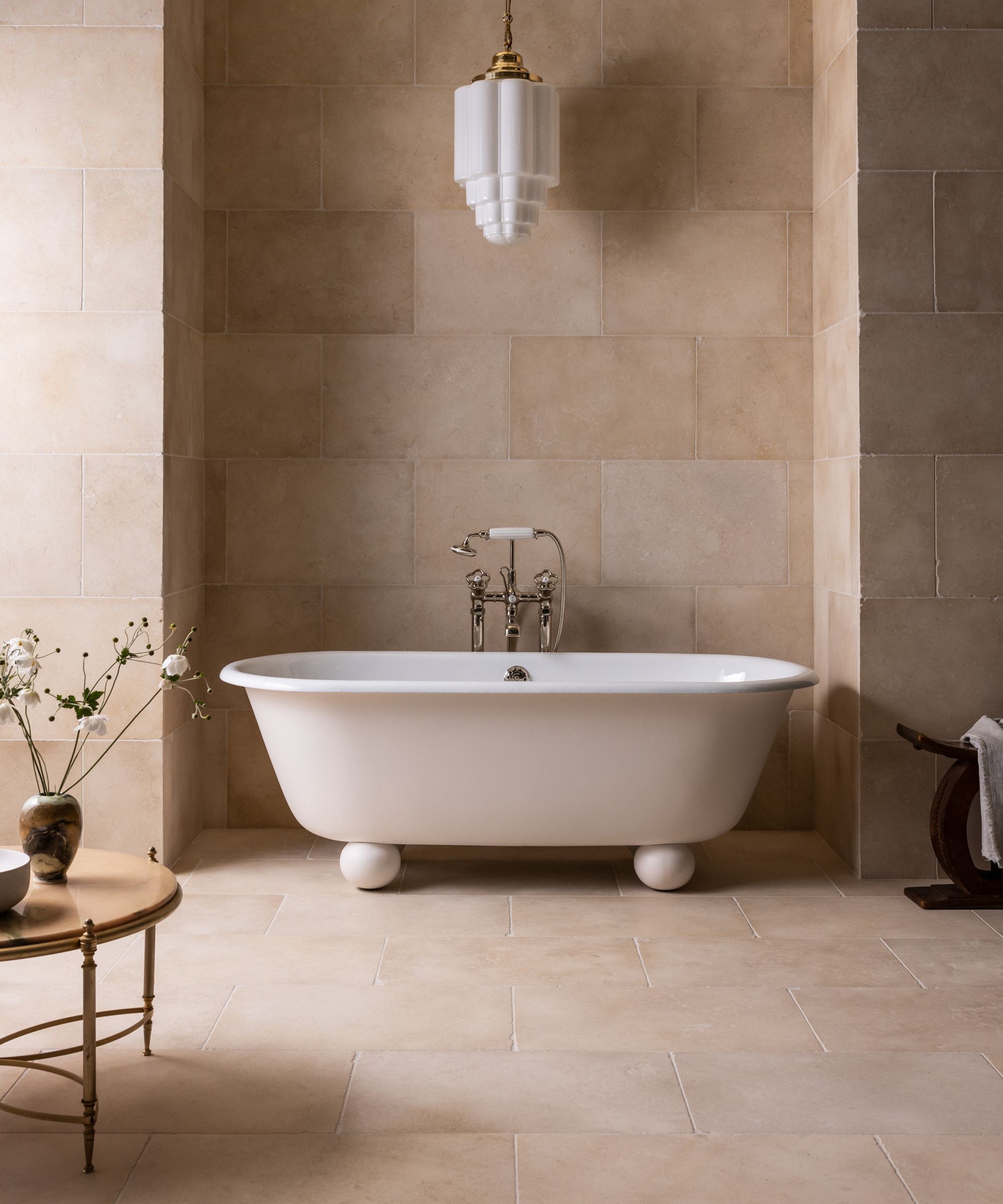 A bathtub with spherical feet, in a room with limestone tiles and a pendant light hanging from the ceiling.
