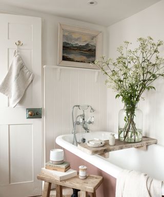 A bathroom with a white tub with a dusky pink base with a bath tray with a vase of wildflowers, a wooden stool next to it with books on, and a white door and mountain wall art print