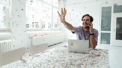 A man celebrates in front of his laptop while confetti blows all around him and across his desk.
