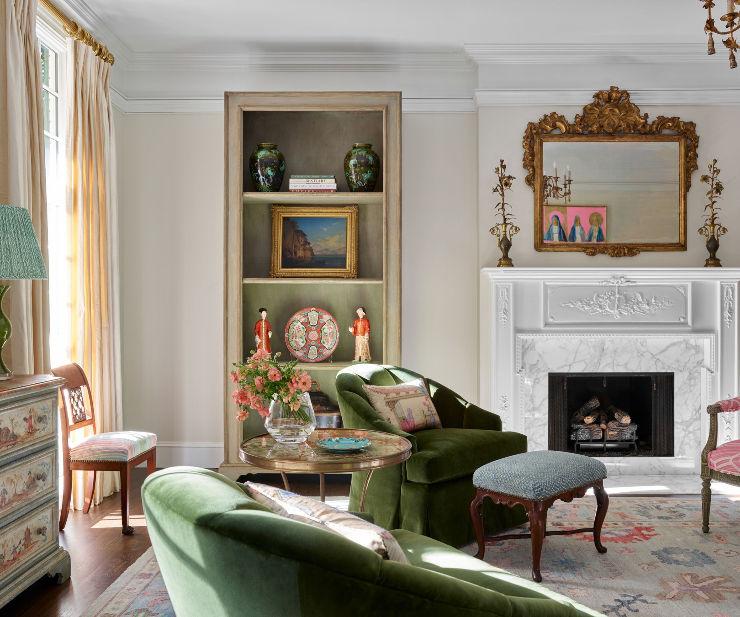 Living room with bookcase of antiques, marble fireplace with antique gold mirror, green velvet armchairs, antique stool and large rug