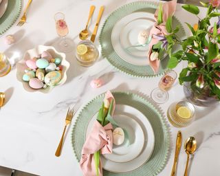 Easter tabletop overhead with gold cutlery, mint green round place mats, white layered dinnerware, fresh florals, and bowl of pastel assorted eggs.