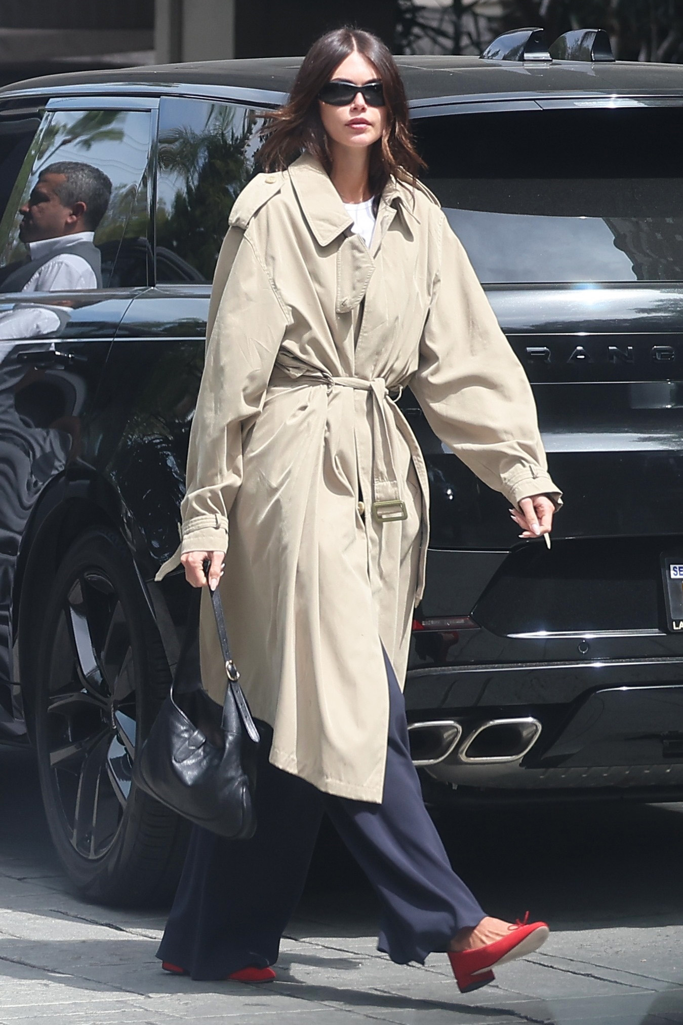 Kaia Gerber wearing a trench coat, a white T-shirt, navy satin pants, and red ballet flats.