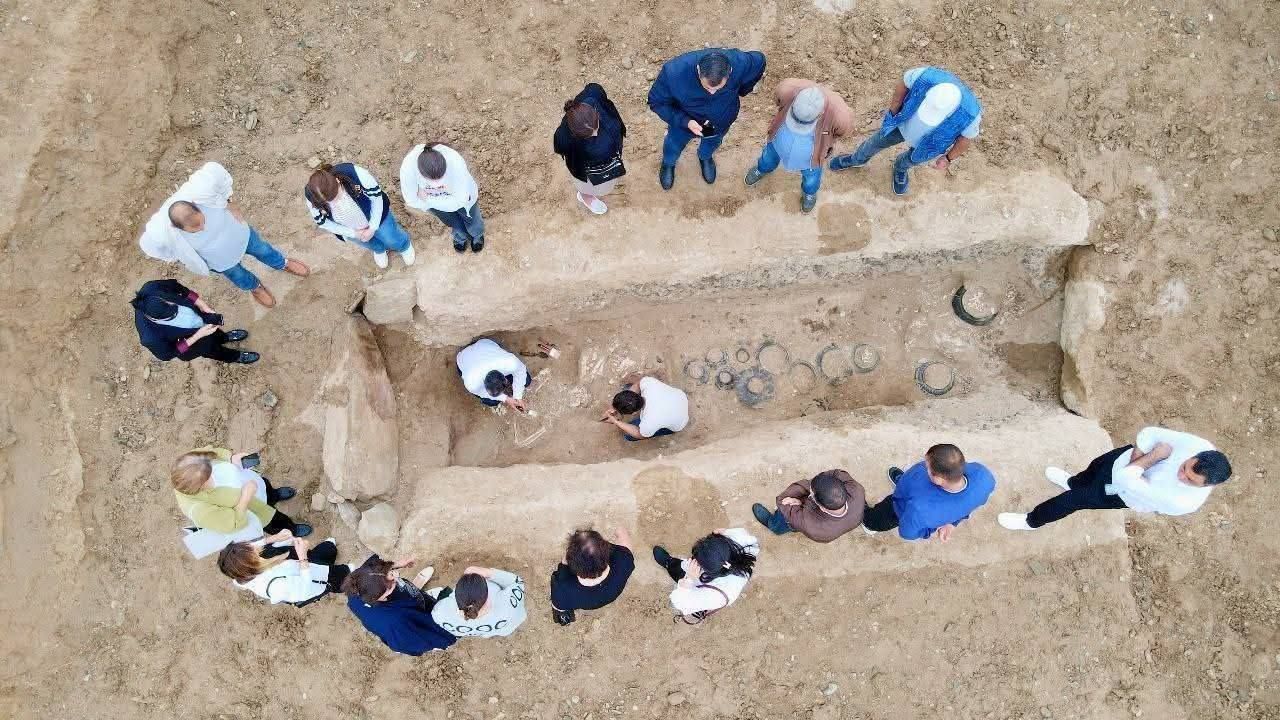 a group of people stand around as two archaeologists excavate a grave