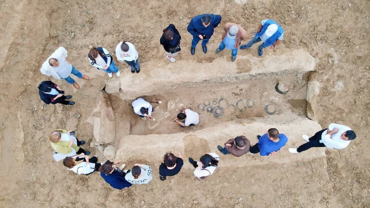 a group of people stand around as two archaeologists excavate a grave