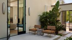 A backyard patio with a large door leading into a living room. The patio features gray paving, a pair of tan chairs, a side table, and lush greenery