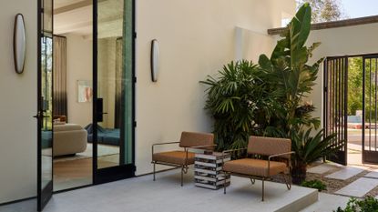 A backyard patio with a large door leading into a living room. The patio features gray paving, a pair of tan chairs, a side table, and lush greenery