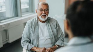 A man with grey hair and beard wearing a white top and shirt sitting in doctors room