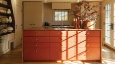 Image of a beige kitchen with french doors letting sunlight in. There is an island with red-orange cabinetry and a matching marble worktop. On the cabinets are round, wooden cabinet knobs. 