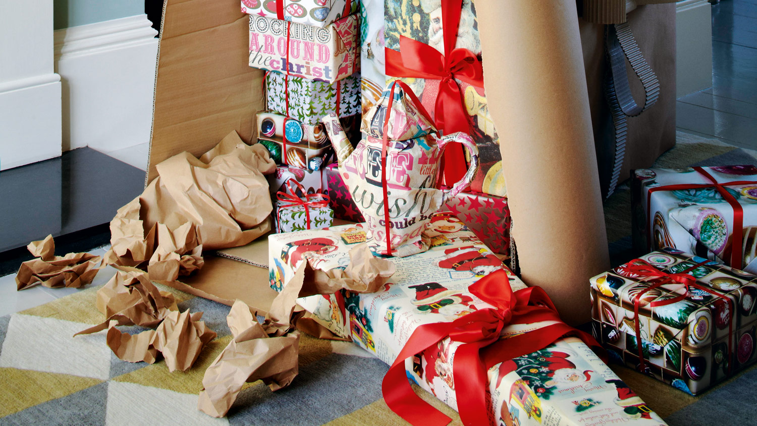 Pile of presents in a large overturned cardboard box, ripped up wrapping paper in from of large box, yellow, cream and grey rug under the presents.