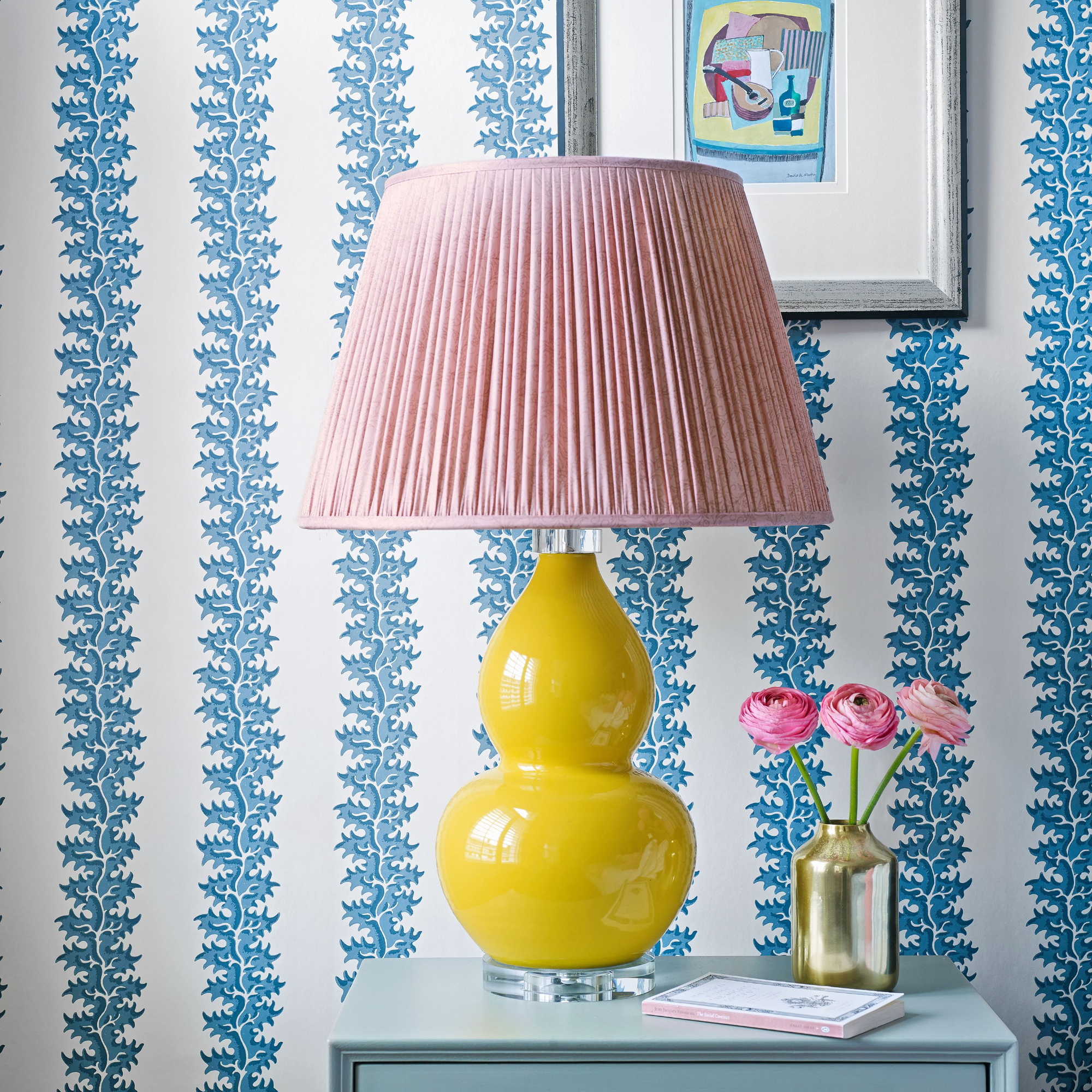 A yellow lamp with pink shade on a bedside table in front of a blue and white wallpapered wall.