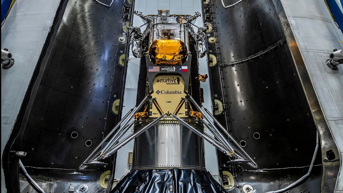 a silver and told spacecraft is seen inside the two black halves of its rocket&#039;s payload fairing in a large, white-walled room