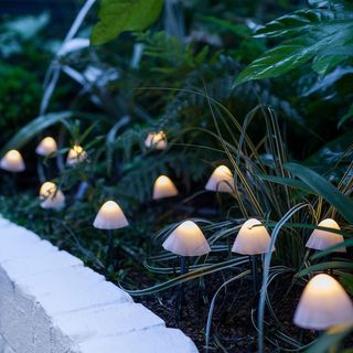 12 Mini Mushroom Solar Stake Lights from Lights4fun, placed in a garden path with a white brick wall