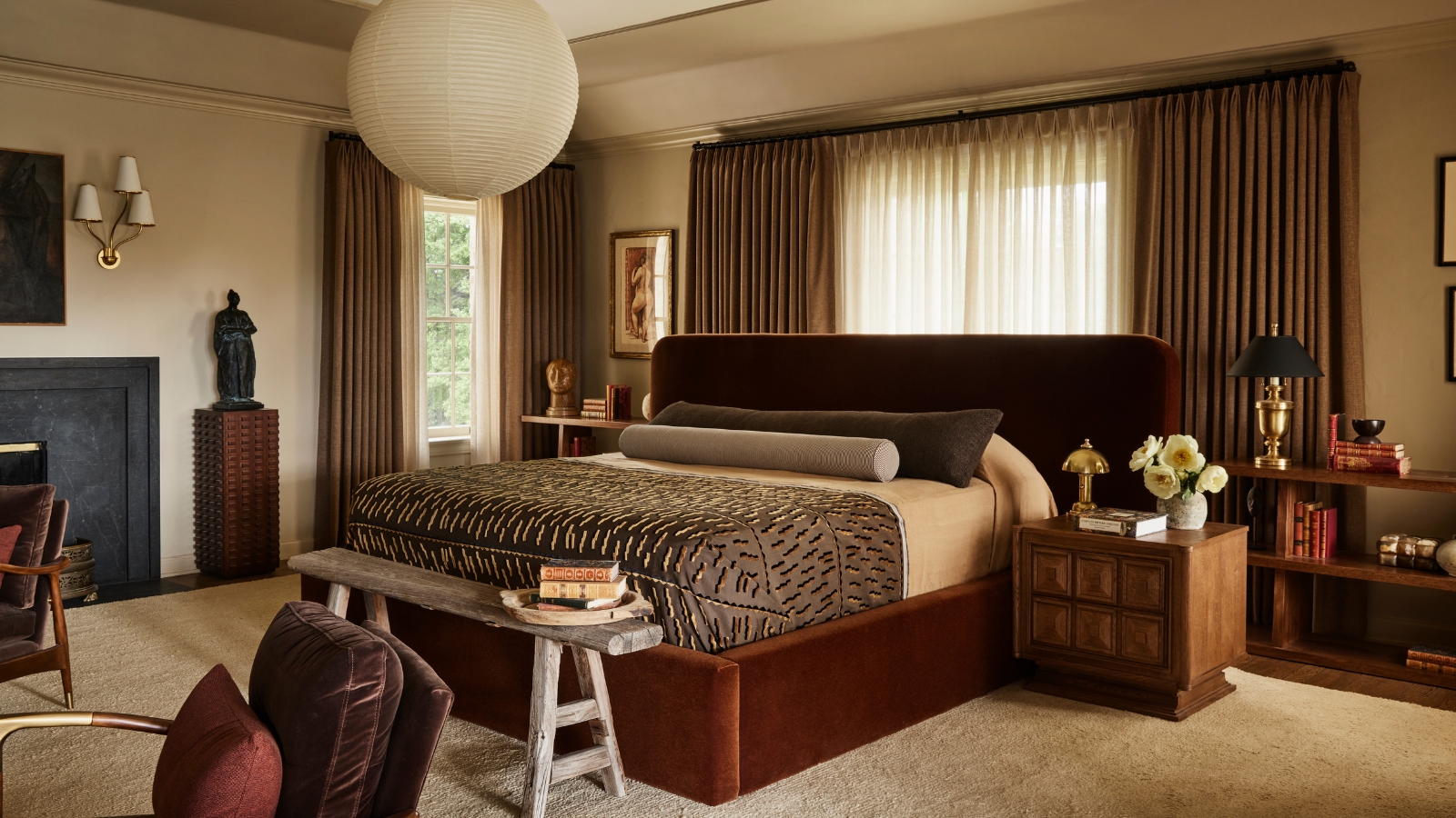 Warm neutral bedroom with a rust velvet bed, brown curtains, wood furniture, patterned coverlet and black fireplace.