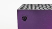Macro shot of the circular Xbox logo power button on the corner of the console, under a purple metallic sheen.