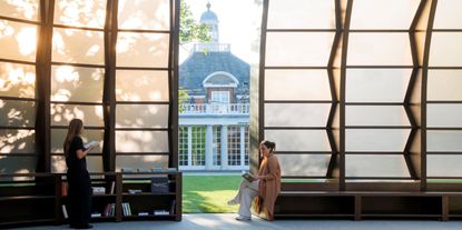 A sun-lit contemporary architecture pavilion sited in a park lets sunshine through its glass and wooden panels as two women spend time in it.