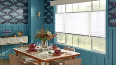 Blue dining room with colorfully set table. Blue walls, floral wallpaper, bright windows, and a flower vase.