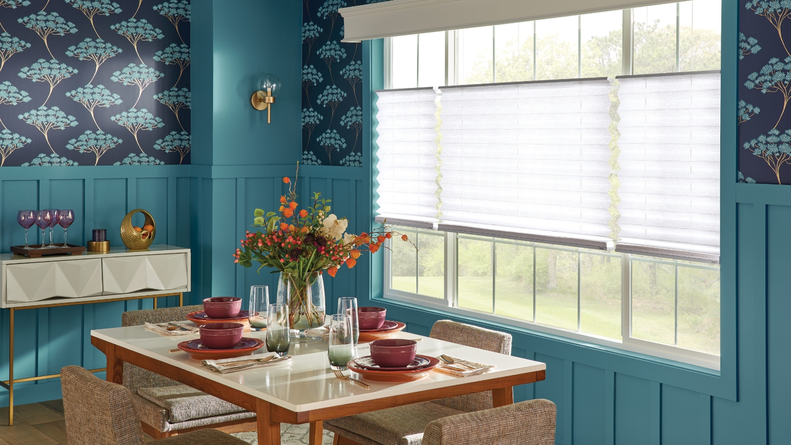 Blue dining room with colorfully set table. Blue walls, floral wallpaper, bright windows, and a flower vase.
