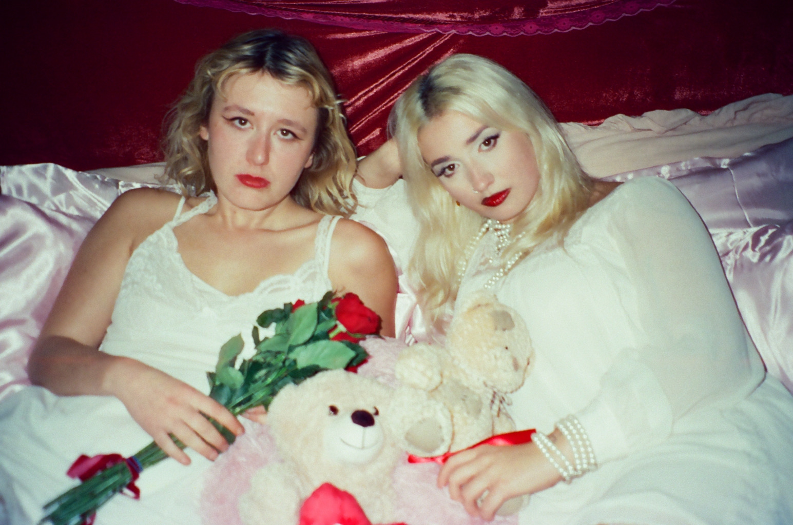 Lambrini Girls in white, holding roses and teddy bears in a bed