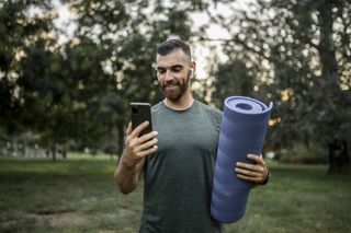 A guy using a fitness app on his phone while holding his yoga mat.