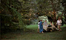 A family stood around a dining table in a woodland with fairy lights strung in the trees