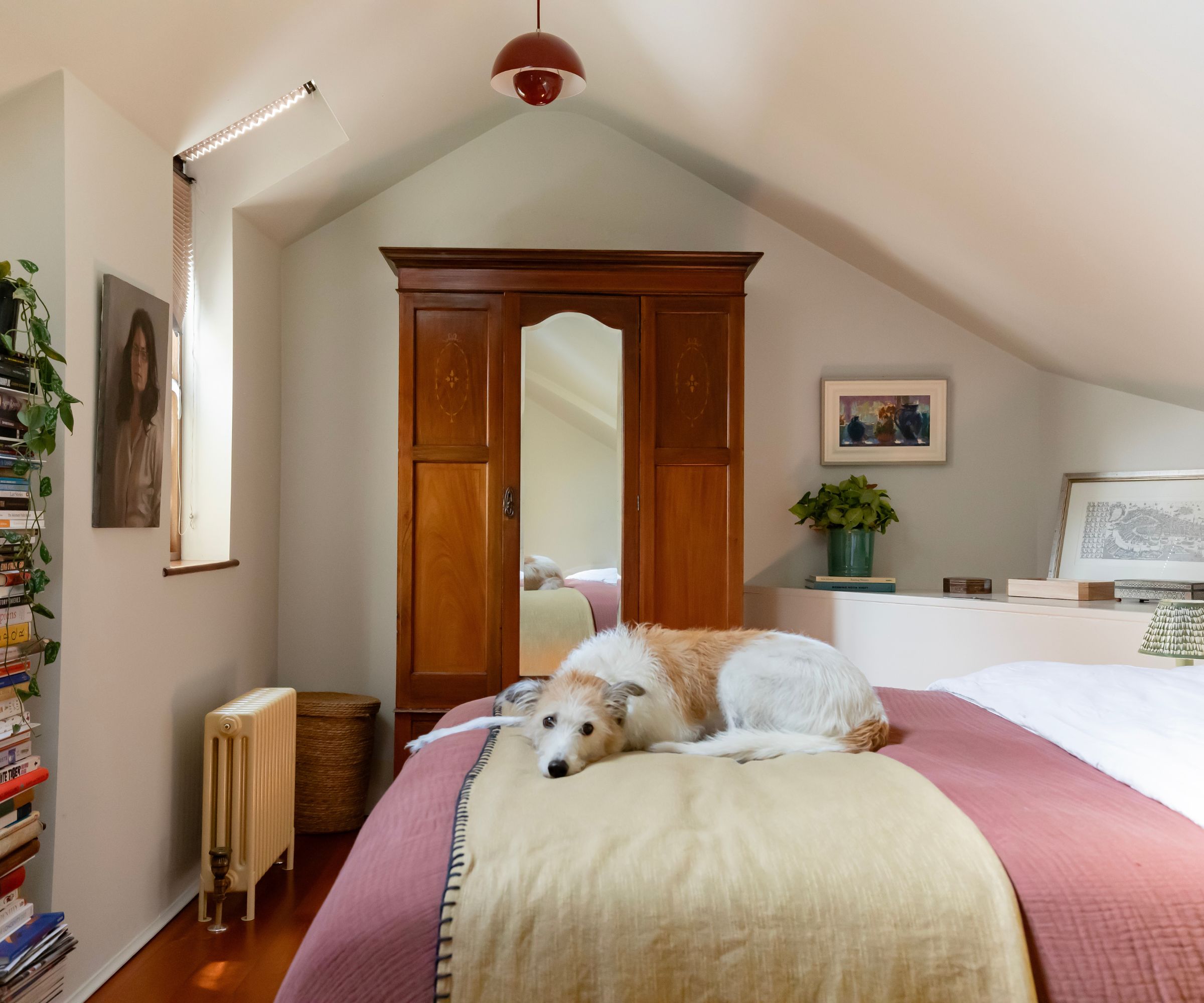 Bedroom with sloped ceiling, white walls, dark wood wardrobe, bed with pink throw and dog