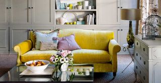 Taupe colour living room with yellow velvet sofa glass coffee table and bespoke built in cupboards to highlight areas where key dusting mistakes might occur