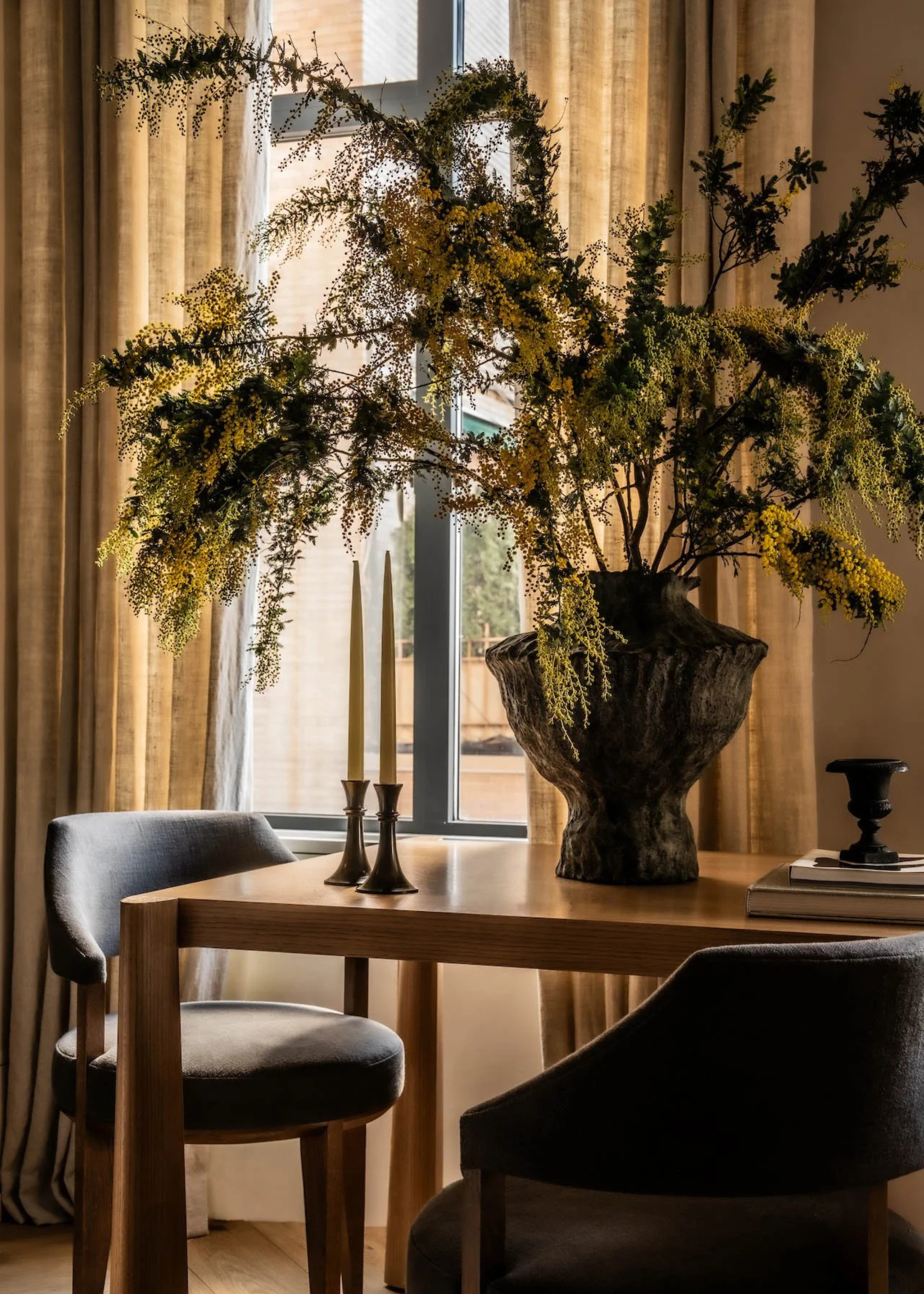 A wood dining table with upholstered chairs by a window with semi-sheer muslin-style curtains, a vase of feathered stems, a pair of candles on holders, and a stack of books