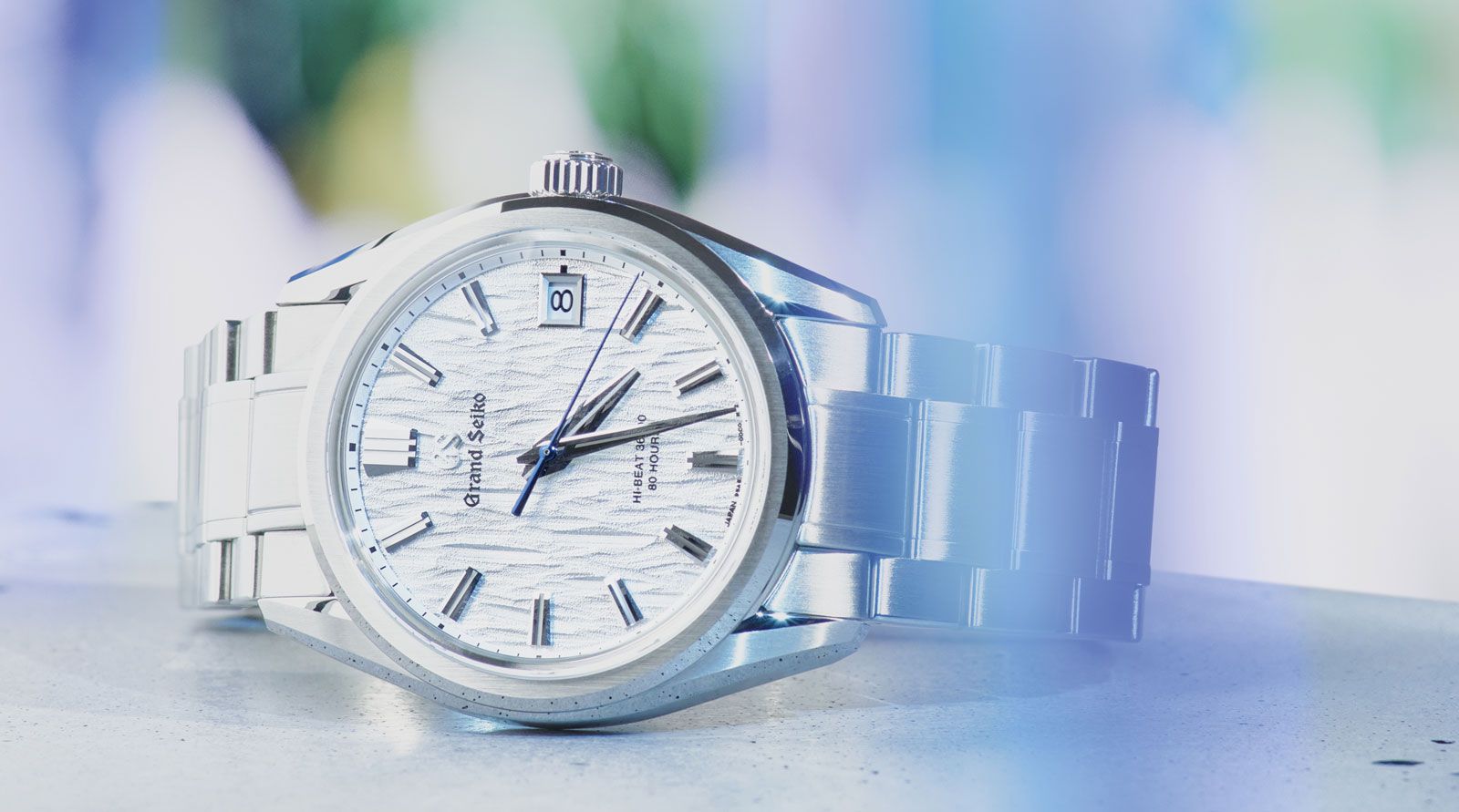 A close-up of a side-ways facing silver coloured Grand Sieko watch. The watch face is silver with a rough edge and the face has Roman numerals.
