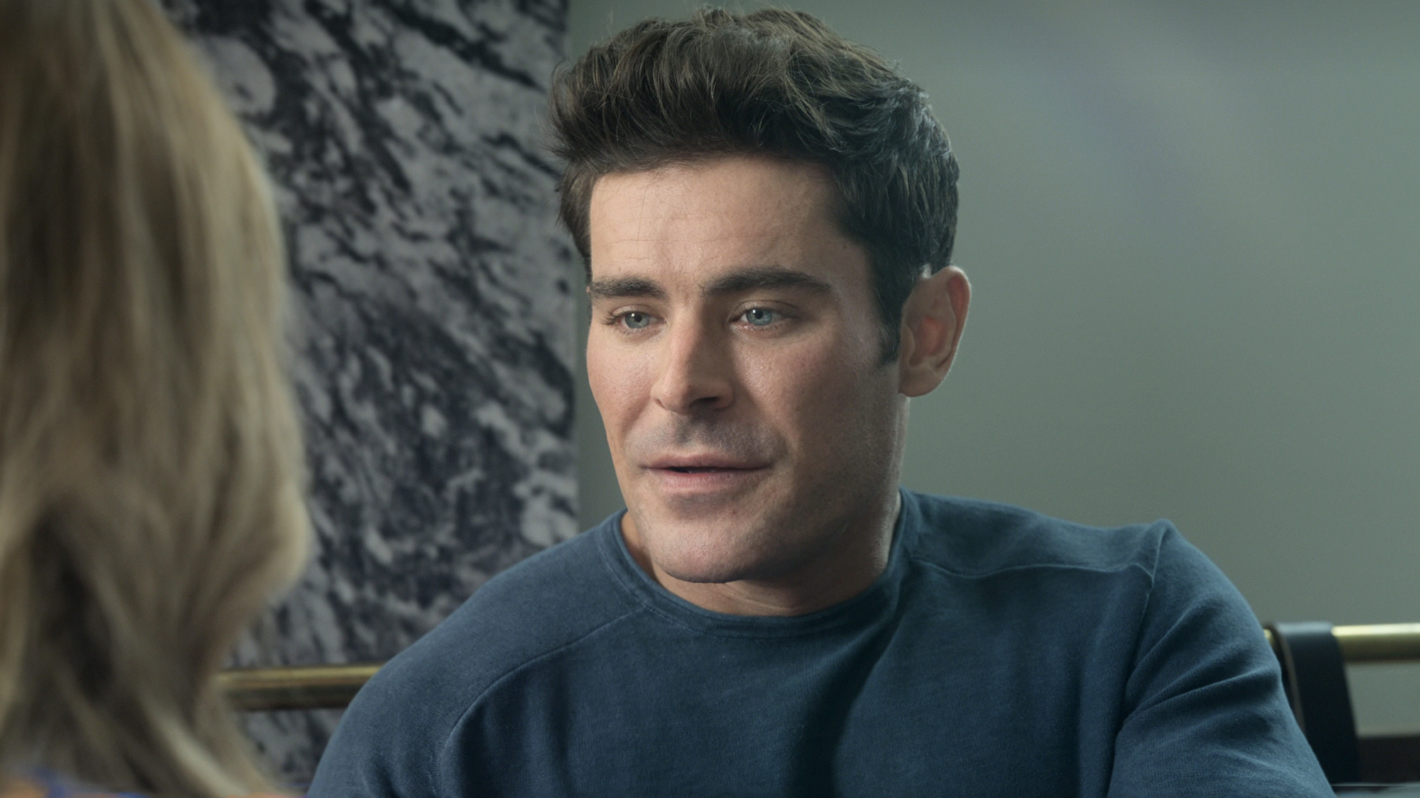 Zac Efron with a blue long sleeve shirt sitting in his starring role opposite Nicole Kidman in Netflix's A Family Affair (2024).