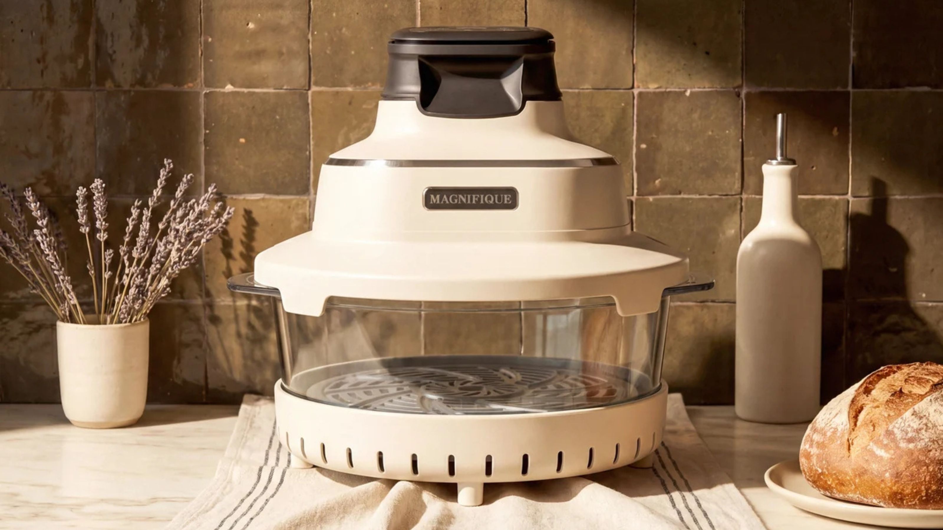 An air fryer with a glass mid section, white base, and white lid on a kitchen counter against a green tile backsplash. 