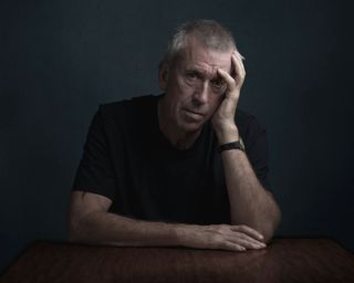 A moody image of an elderly individual with arms on the table looking at the camera, on a dark backdrop.