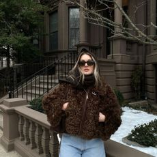 A woman wrapped up in a brown fluffy coat standing outside a snowy brown house surrounded by trees