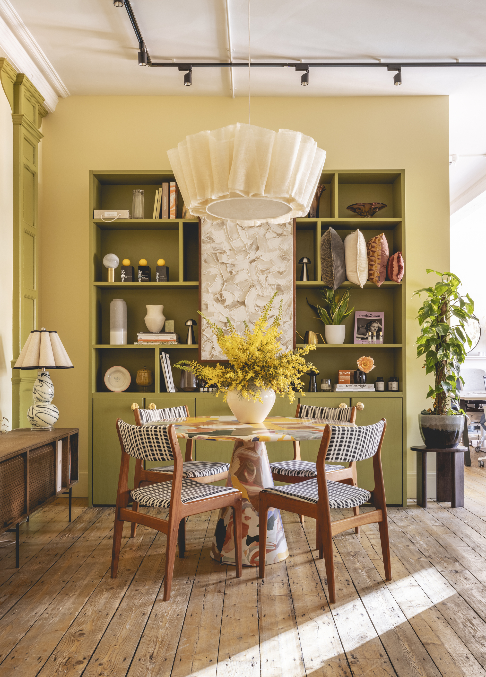 dining room with yellow walls, green bookshelf styled with decor, round table with upholstered dining chairs, a vase of yellow flowers on the table with a pendant lamp above, and a timber console table