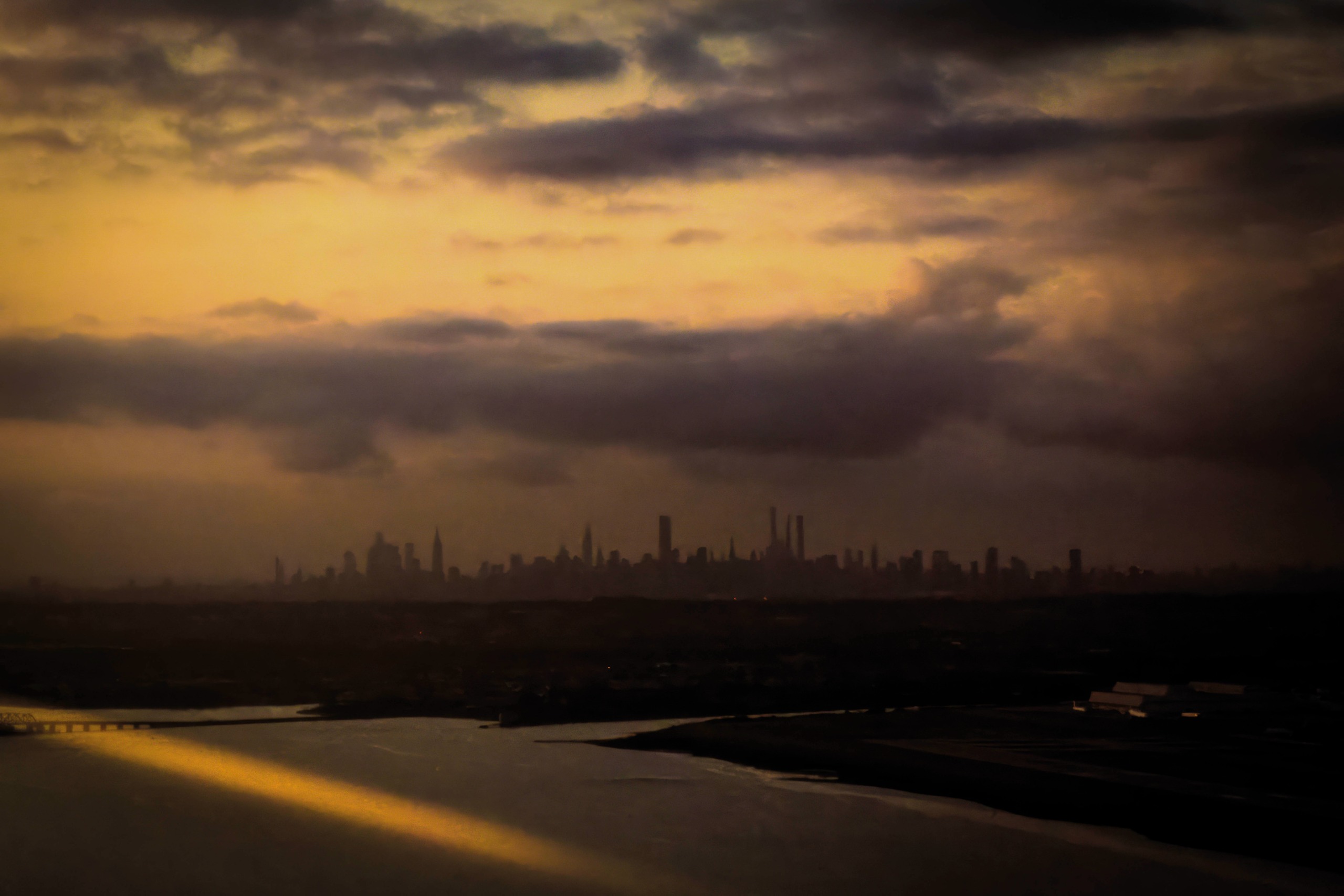 The Manhattan skyline appears as a faint dark silhouette beneath heavy storm clouds tinted amber by the setting sun.