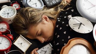 A woman with blonde hair sleeps surrounded by alarm clocks