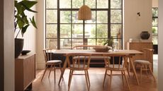 A large wooden dining table surrounded by six matching wooden chairs with gray cushions on them. A hanging rattan pendant light above, and a large floor to ceiling lead window behind. Wooden herringbone floor