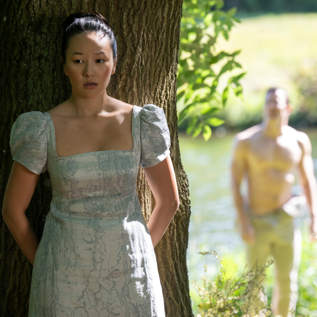 A Regency-era woman (Yerin Ha as Sophie Baek) hides behind a tree as a shirtless man (Luke Thompson as Benedict Bridgerton) stands blurred in the background, in front of a lake, in episode 403 of Bridgerton.