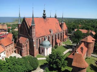 a red brick cathedral stands in a grassy area surrounded by like buildings. Green trees stretch to the horizon.