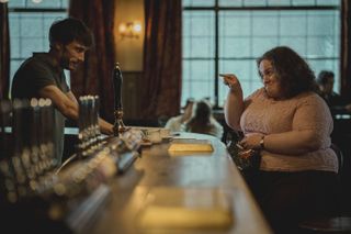 a male barkeep (Richard Gadd as Donny) stands behind the bar as a female patron (Jessica Gunning as Martha) points at him and smiles, in the netflix series 'baby reindeer'