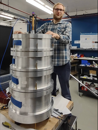 a man in a blue flannel shirt stands behind a tall silver cylinder in a laboratory