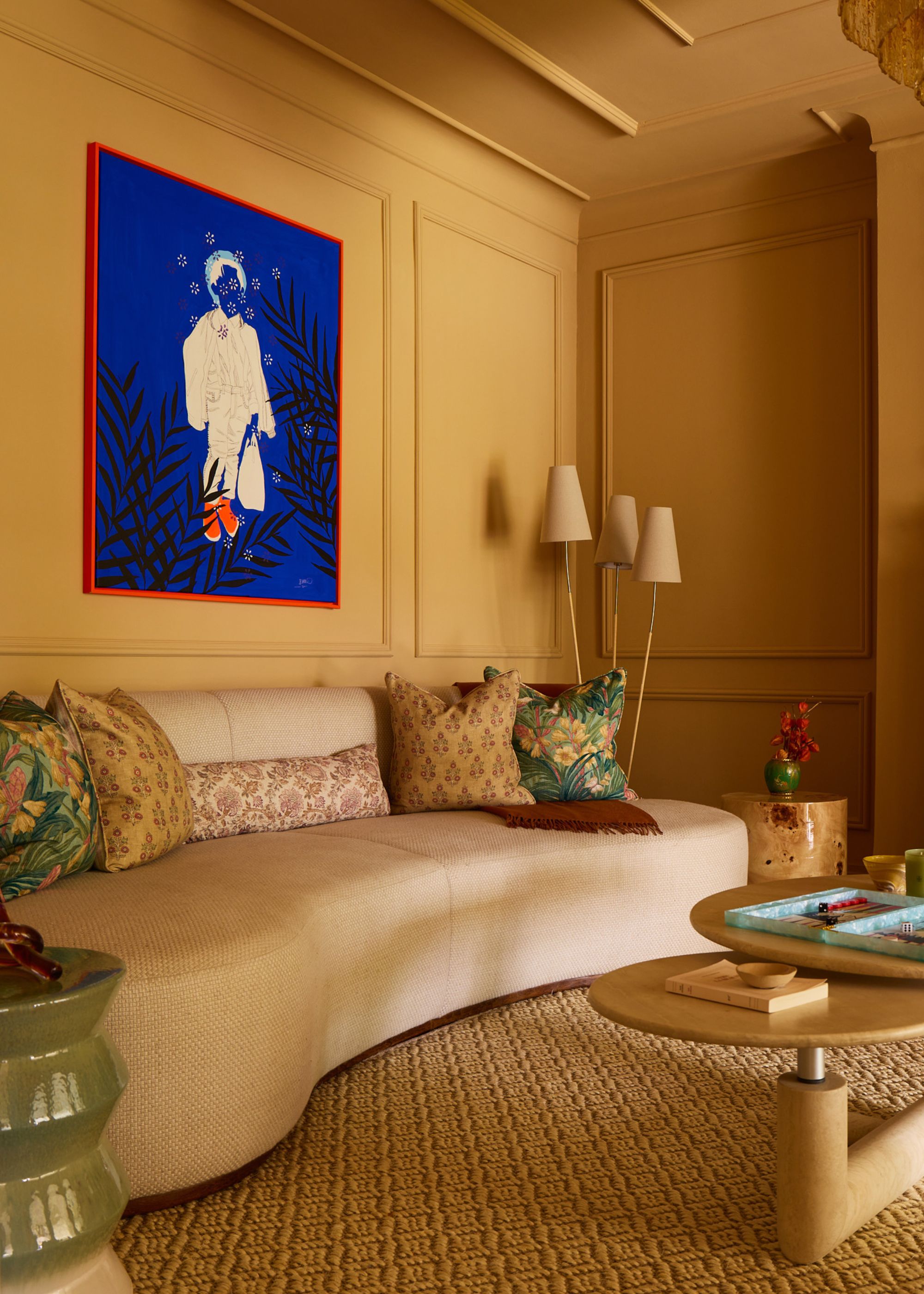 A large living room with traditional wall and ceiling panelling, color-drenched a dark yellow. A modern, sculptural cream sofa with patterned accent cushions, and an electric blue artwork on the wall.