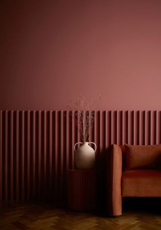 A room painted in a pinky red with an orange sofa and wooden herringbone flooring