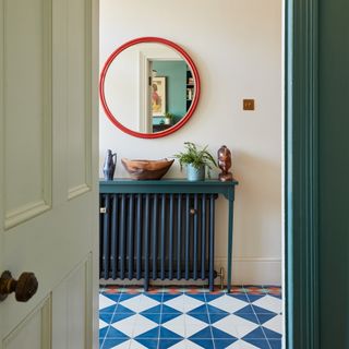 view into a hallway with a tiled floor in blue, white and red with a console table over a blue painted radiator and a red framed round mirror